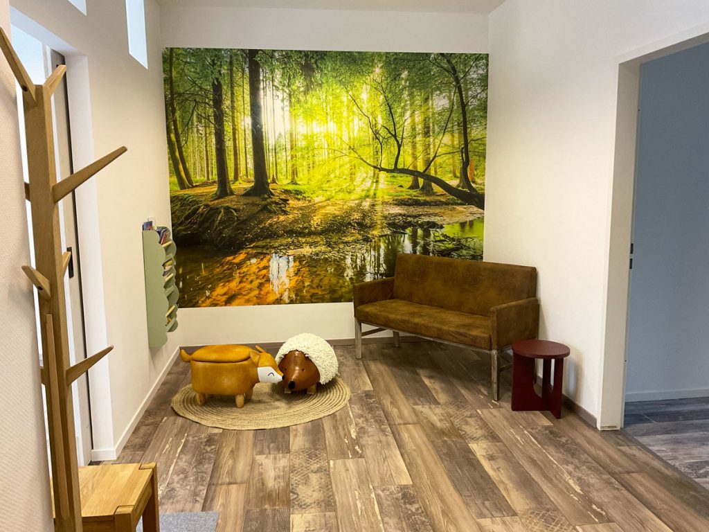 Zeigt den Eingangsbereich der Physiotherapie. Zu sehen ist ein großes Wald-Wandbild, eine braune gemütliche Couch und zwei Sitzhocker in Form eines Igels und eines Fuchses.