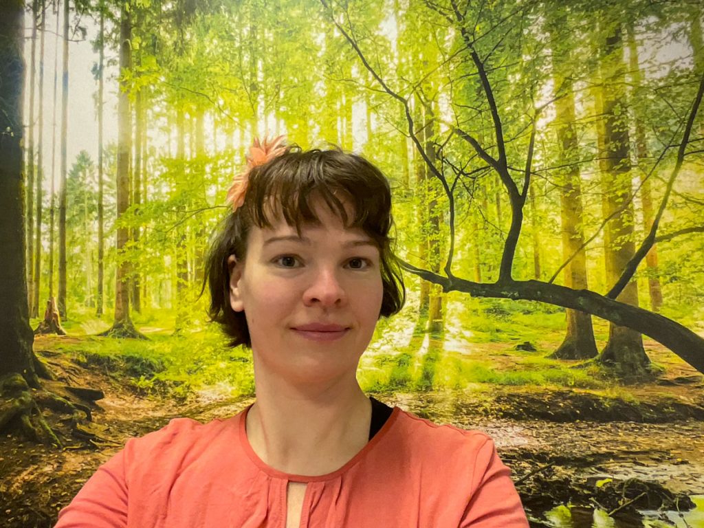 Foto von Stefanie Venzke. Sie lächelt freundlich, hat eine Blume im Haar und steht vor einem Waldhintergrund-Bild