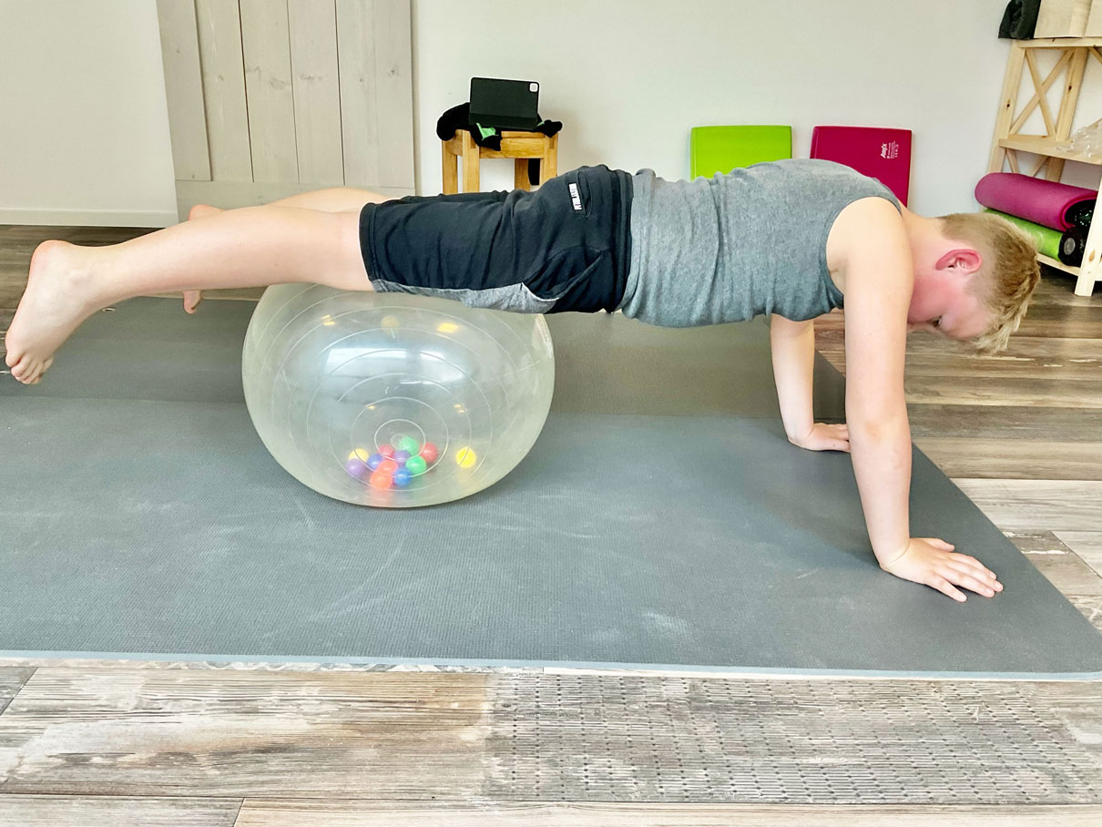 Junge macht Gymnastik. Dabei liegen seine Beine auf einen großen aufgeblasenen Ball und seine Arme sind auf dem Boden aufgestützt.
