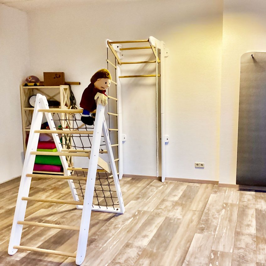 Zeigt den einladenden Praxisbereich für Kinder. Es gibt ein Klettergerüst, eine Handpuppe, Bälle und weiteres Spielzeug zur Förderung der Bewegung.